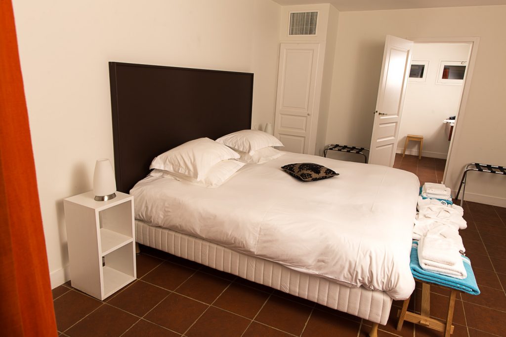 Grande chambre aux murs blancs avec un lit double entouré de tables de chevet et d’une salle d’eau dans le fond
